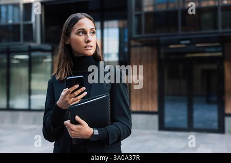 Selbstbewusste und seriöse Frau in schwarzer Kleidung, die ein Smartphone und ein digitales Tablet vor einem Glasgebäude hält. Junge Geschäftsfrau trägt Stockfoto