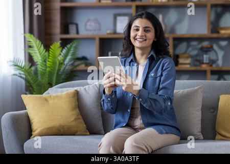 Eine junge Indianerin lächelt, während sie ihr Tablet zu Hause auf der Couch in ihrem Wohnzimmer benutzt; sie trägt ein blaues Jeanshemd. Stockfoto