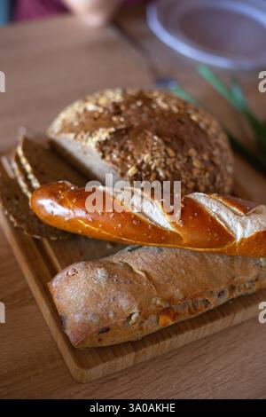 Eine Auswahl an frisch gebackenem Brot, darunter ein rustikaler Vollkornbrot, eine weiche Brezel und ein Baguette mit Sättigung auf einem Holzbrett. Krieg Stockfoto