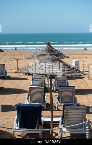 An einem marokkanischen Strand mit Meerblick stehen Liegestühle unter Strohschirmen. Stockfoto