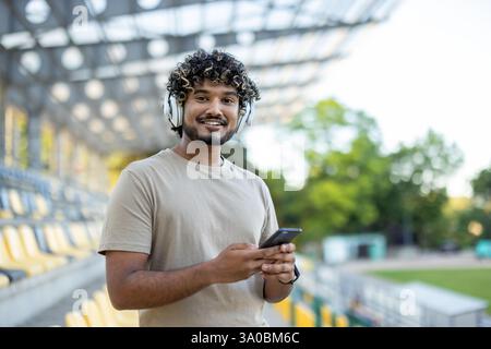 Sportler, der mit dem Handy in der Hand in die Kamera schaut, Mann, der beim Joggen Kopfhörer trägt, aktive körperliche Übungen macht. Stockfoto