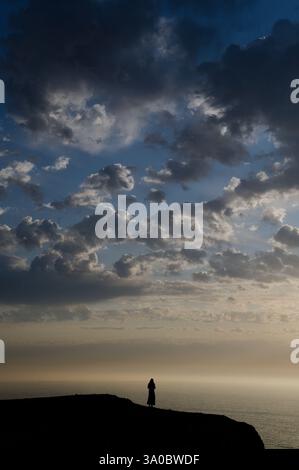 Eine Einzelfigur steht auf einer Küstenklippe unter einem dramatischen Himmel bei Sonnenuntergang am Tonel Beach Stockfoto