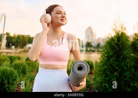 Die fröhliche junge Frau in sportlicher Kleidung bereitet sich auf Yoga in üppigem Grün in einem urbanen Park vor. Stockfoto