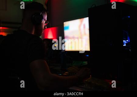 Junger Mann mit Brille und Kopfhörern, der in einem Computerspielzimmer sitzt und sich auf einen Bildschirm konzentriert, mit farbenfrohen Neonlichtern im Hintergrund. Stockfoto