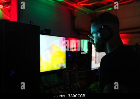 Junger Mann mit Brille und Kopfhörern, der in einem Computerspielzimmer sitzt und sich auf einen Bildschirm konzentriert, mit farbenfrohen Neonlichtern im Hintergrund. Stockfoto