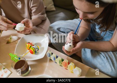 Ein Mädchen malt schwarze Augen auf ein weißes Osterei, während es vorsichtig hält. Auf dem Tisch stehen bunte Farben und Pinsel. Zwei dekorierte Eier wi Stockfoto