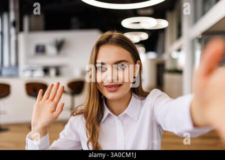 Lächelnde Frau, die während eines Videogesprächs im modernen Büro winkt. Freundliche Geste schafft einladende Interaktionen. Professionelles Ambiente mit Multimedia-Communi Stockfoto