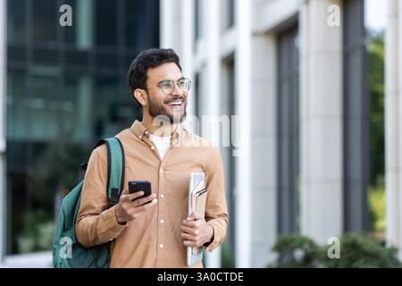 Junger Mann, der mit einem Rucksack durch die Stadt läuft, Student lächelt vor dem Universitätsgelände in lässiger Kleidung. Stockfoto