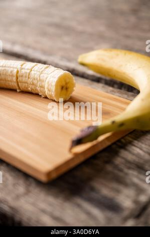 Eine teilweise geschnittene Banane liegt auf einem hölzernen Schneidebrett, dessen Schale in der Nähe liegt, vor einem rustikalen Holztischhintergrund. Stockfoto