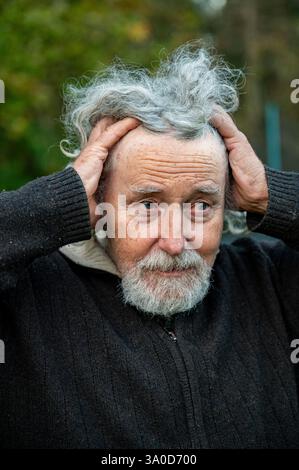Ein älterer Mann mit lockigen grauen Haaren und Bart hält mit beiden Händen den Kopf und sieht besorgt aus. Er trägt einen dunklen Pullover und der Hintergrund ist unscharf Stockfoto