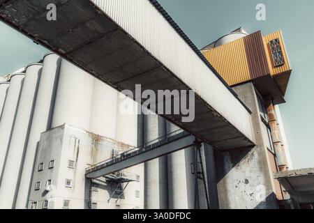 Weitwinkelaufnahme einer modernen Industrieanlage mit großen zylindrischen Kornsilos, einer verwitterten Betonkonstruktion und einem erhöhten geschlossenen Gehweg c Stockfoto