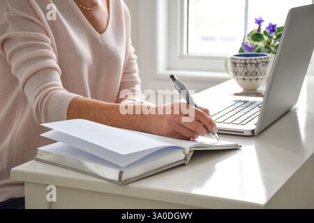 Frau, die an einem Schreibtisch sitzt, mit Laptop-Computer, der Notizen in Notizbuch schreibt Stockfoto