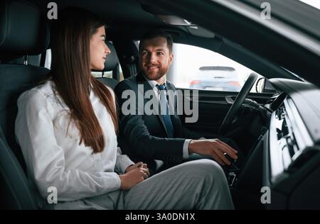 Ein Mann in Anzug und Krawatte sitzt mit einer jungen Frau, einem Kunden, im Auto und zeigt den Innenraum des Autos. Stockfoto