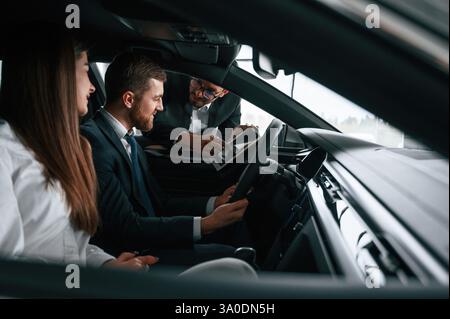 Ein Mitarbeiter im Autohaus berät Mann und Frau, die im Auto sitzen. Stockfoto