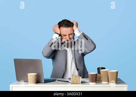 Junger Firmenarbeiter jammerte oder weinte, packte verzweifelt seinen Kopf und saß am Schreibtisch auf blauem Studiohintergrund Stockfoto