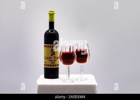 Eine Miniatur-Weinflasche mit zwei gefüllten Gläsern auf einem weißen Sockel. Diese Makroaufnahme erfasst feine Details und erzeugt die Illusion eines echten Weins Stockfoto