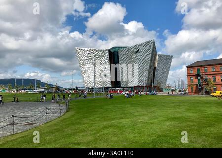 Titanic Belfast Museum an einem sonnigen Tag mit seiner beeindruckenden modernen Architektur und einem lebhaften grünen Park, in dem Besucher die Umgebung genießen. Stockfoto