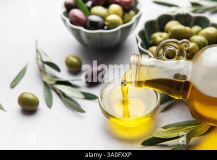 Goldenes Olivenöl, das aus einer Glasflasche in eine kleine Schüssel gefüllt wird, umgeben von frischen Oliven und Olivenzweigen Stockfoto