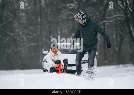 Glückliche Paare sind im Winter draußen. Der Mann nimmt seine Frau auf einen Schlitten. Weißes Auto hinten. Stockfoto