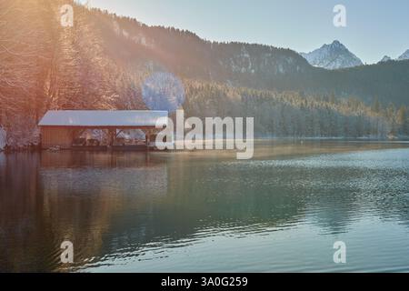 Dieses hochauflösende Foto fängt einen atemberaubenden Wintersonnenaufgang an einem ruhigen Seeufer ein. Der Schwerpunkt ist ein rustikales hölzernes Bootshaus, teilweise s Stockfoto
