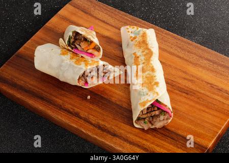 Köstliche gegrillte Rindfleisch-Shawarma-Wrap mit frischem Gemüse, serviert auf einem Holzbrett, isoliert auf dunklem Hintergrund Stockfoto