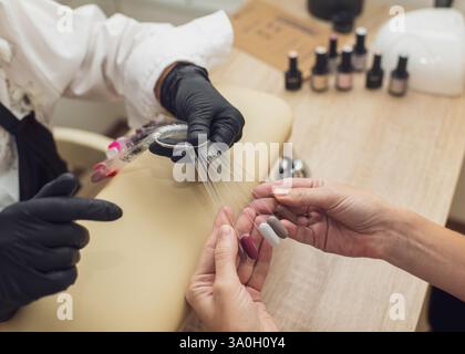Ein Maniküre in schwarzen Handschuhen wählt mit einem Kunden ein Nageldesign, bevor er eine Maniküre mit einer Fächerpalette durchführt. Stockfoto