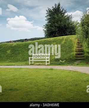 Weiße Bank in einem Park. Weiße Holzbank in einem landschaftlich gestalteten Park in der Nähe eines kleinen Hügels. Zwei Radfahrer fahren im Hintergrund auf dem Hügel. Stockfoto