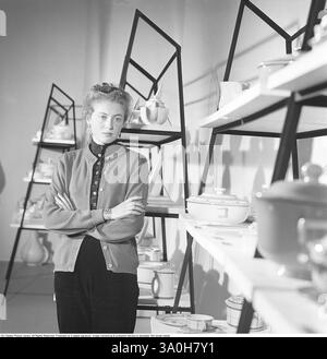Frau in den 1950ern Eine Frau, die selbstbewusst mit überkreuzten Armen vor einer Ausstellung moderner Küchenutensilien aus der Mitte des Jahrhunderts steht. Sie trägt eine stilvolle, geknöpfte Strickjacke über einem Hemd mit Kragen, kombiniert mit einem Rock, der die Mode dieser Zeit widerspiegelt. Ihr Haar ist ordentlich gestylt in einer eleganten Hochsteckfrisur, typisch für die 1950er Jahre Hinter ihr befinden sich elegante, geometrische Regale, in denen eine Auswahl an Keramikgeschirr, Teekannen, Schüsseln und anderen Haushaltsgegenständen aufbewahrt wird, die das klare, funktionale skandinavische Design der Mitte des 20. Jahrhunderts zeigen. ref. BV85-1 Stockfoto