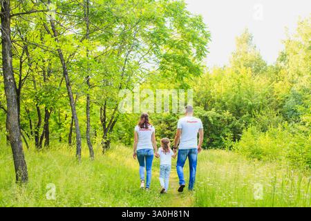 Dieses herzerwärmende Foto zeigt eine dreiköpfige Familie, die Hand in Hand durch einen üppigen grünen Wald geht. Das helle und lebhafte Laub umgibt sie, giv Stockfoto