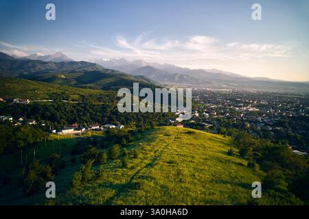 Panoramablick auf die Drohne mit grünem Hügel und Häusern mit Landschaft der schneebedeckten Berge im Hintergrund in Almaty Stadt, Kasachstan Stockfoto