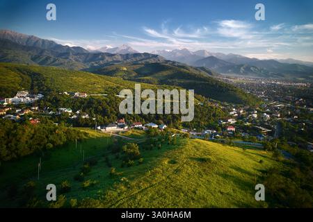 Panoramablick auf die Drohne mit grünem Hügel und Häusern mit Landschaft der schneebedeckten Berge im Hintergrund in Almaty Stadt, Kasachstan Stockfoto