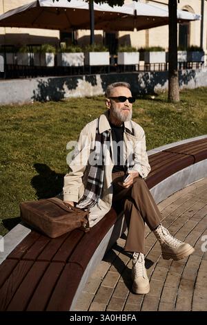 In einem belebten Stadtpark sitzt ein gutaussehender Seniorenmann in trendiger Kleidung auf einer Holzbank. Stockfoto