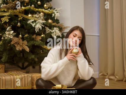 Eine süße junge Frau sitzt vor einem Weihnachtsbaum und goldenen Schachteln mit Geschenken Stockfoto