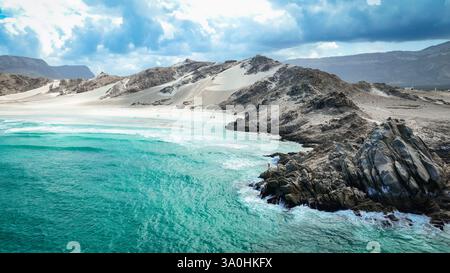 Wellen prallen sanft gegen zerklüftete Felsen, während Sanddünen hinter dem ruhigen türkisfarbenen Wasser an der Küste von Socotra aufsteigen. Stockfoto