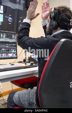 Ein Fernsehregisseur in einem Senderaum, der aktiv eine Live-Produktion leitet. Umgeben von mehreren Bildschirmen trägt der Regisseur ein Headset und bewegt sich in Richtung eines Video-Umschalters. Stockfoto