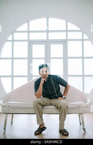 Ein nachdenklicher Mann ruht sich auf einem stilvollen Sofa aus, von Hand bis zum Kinn. Entspannen Sie sich in einem hellen, modernen Zimmer, die Atmosphäre fördert Nachsicht und Ruhe. Idee Stockfoto