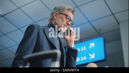 Überfüllter internationaler Flughafenterminal: Reifer Mann hält Flugticket, telefoniert mit dem Mobiltelefon, um Fluginformationen zu überprüfen. Reisende, die auf Urlaubsreise gehen. Im Hintergrund laufen verschiedene Menschen. Stockfoto