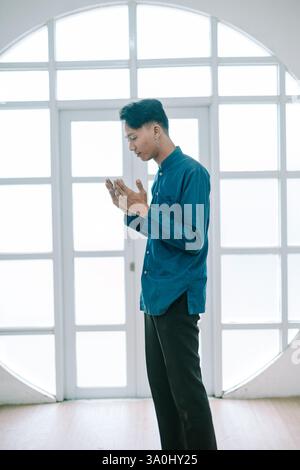 Ein besinnlicher junger Mann asiatischer Herkunft steht im Gebet vor einem Fenster. Er trägt ein blaues Hemd und eine dunkle Hose, Hände in einer Geste von Devoti gehalten Stockfoto