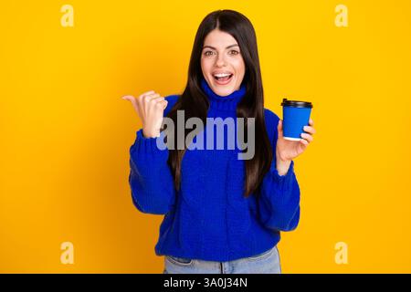 Lächelnde Frau in blauem Pullover, der Kaffeetasse hält und Gesten auf leuchtendem gelbem Hintergrund macht Stockfoto