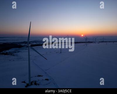 Luftaufnahme eines Windparks in einer schneebedeckten Landschaft bei Sonnenuntergang. Windturbinen stehen hoch gegen das warme Leuchten der untergehenden Sonne und erzeugen einen Stockfoto