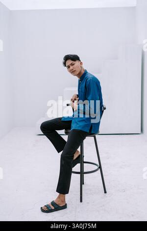Ein junger asiatischer Mann sitzt lässig auf einem minimalistischen Hocker. Er trägt modische Kleidung und zeigt eine nachdenkliche Pose und blickt selbstbewusst in die Stockfoto