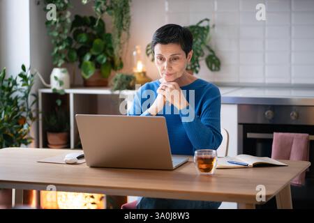 Nachdenklich engagierte, reife Frau lehnt sich auf die Hände, arbeitet an einem Laptop, sieht Seminare an, surft im Internet Stockfoto