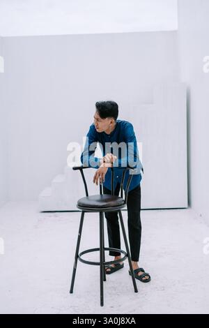 Ein besinnlicher junger Mann in lässiger Kleidung lehnt sich in einem minimalistischen weißen Studio an einen Stuhl. Seine Pose deutet auf ein nachdenkliches und reflektierendes m hin Stockfoto
