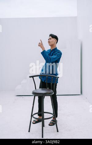 Ein nachdenklicher junger Mann in stilvoller Freizeitkleidung zeigt in einem minimalistischen weißen Studio-Setting nach oben. Die Szene weckt ein Gefühl der Besinnung, indirekt Stockfoto