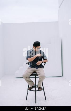 Ein besinnlicher Mann mit Sonnenbrille und lässiger Kleidung sitzt auf einem schwarzen Stuhl in einem minimalistischen weißen Raum. Die Szene weckt ein Gefühl von Ruhe und Moderne Stockfoto