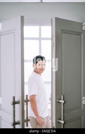 Porträt eines Mannes in lässiger Kleidung, der zwischen offenen Türen in einem hellen, modernen Interieur steht. Er blickt direkt auf den Zuschauer mit einem Co Stockfoto
