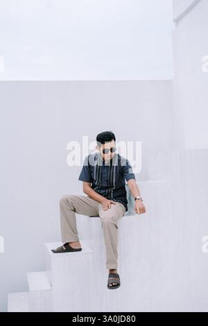 Ein junger Mann sitzt stilvoll auf einer sauberen, weißen, minimalistischen Treppe. Er trägt eine Sonnenbrille und ein lässiges Outfit, das einen modernen und trendigen Look schafft. Perfekt Stockfoto