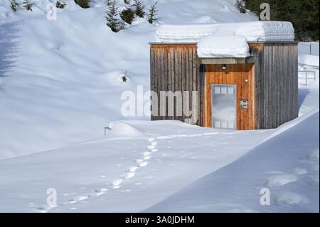 Kleines Wasserkraftwerk, das im Winter tief verschneit ist. Holzgebäude, isoliert in einer riesigen, schneebedeckten Landschaft mit menschlichen Fußspuren. Stockfoto