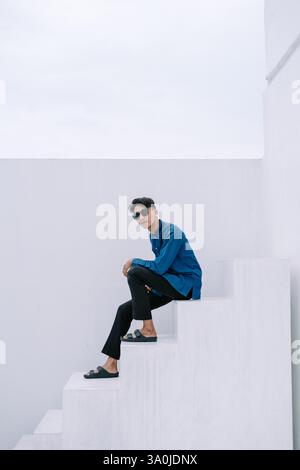 Ein trendiger junger Mann in blauem Hemd und schwarzer Hose sitzt lässig auf weißen Stufen. Sonnenbrillen verleihen seinem modernen Stil einen coolen Touch. Dieses Bild wird aufgenommen Stockfoto