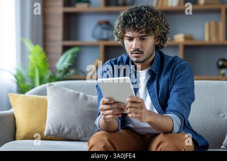 Ein junger Mann mit lockigen Haaren sieht verwirrt aus, während er in einem modernen Wohnzimmer auf seinem Tablet stöbert. Er scheint von etwas überrascht zu sein, das er sieht. Stockfoto